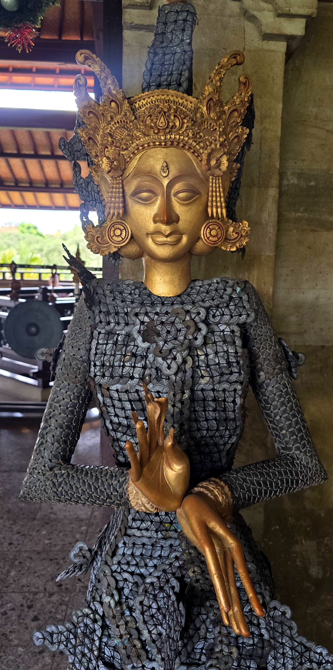 Ornate gold-faced Balinese-style statue in a temple, wearing dark woven coin armor and raised golden hands, with a serene expression.