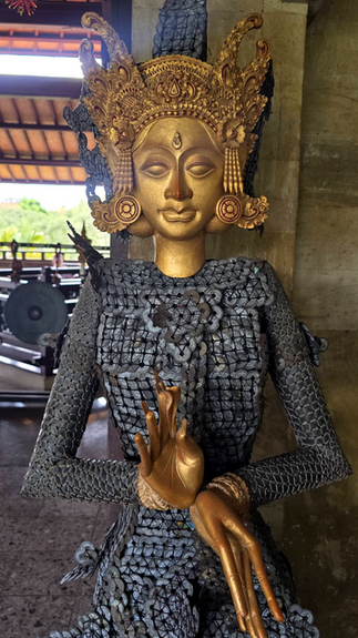 Ornate gold-faced Balinese-style statue in a temple, wearing dark woven coin armor and raised golden hands, with a serene expression.