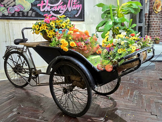 A vintage tricycle loaded with colourful flowers in woven baskets, set on a wooden floor. A sign with "thang Nam" is in the background.