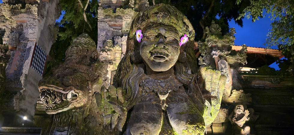 Stone statue with intricate carvings, adorned with flowers, in a temple setting. Lush greenery and twilight sky in the background.