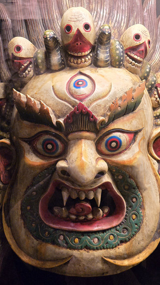 Fierce mask with bulging eyes and fangs, adorned with multiple small faces and intricate patterns, conveying a dramatic and intense mood.