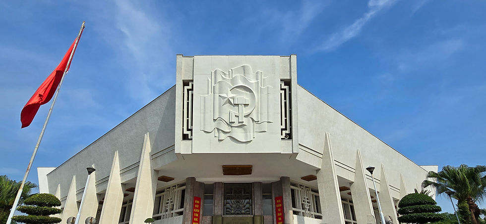 Modern white building with artistic facade and red flag, under clear blue sky. Tropical plants and red banners in front, creating a solemn mood.