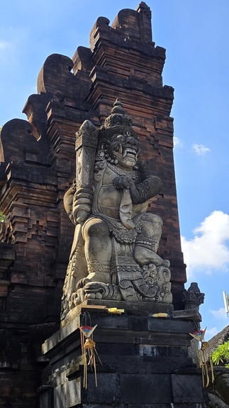 Stone guardian statue at a temple gate, holding a club, against a bright blue sky with offerings below.