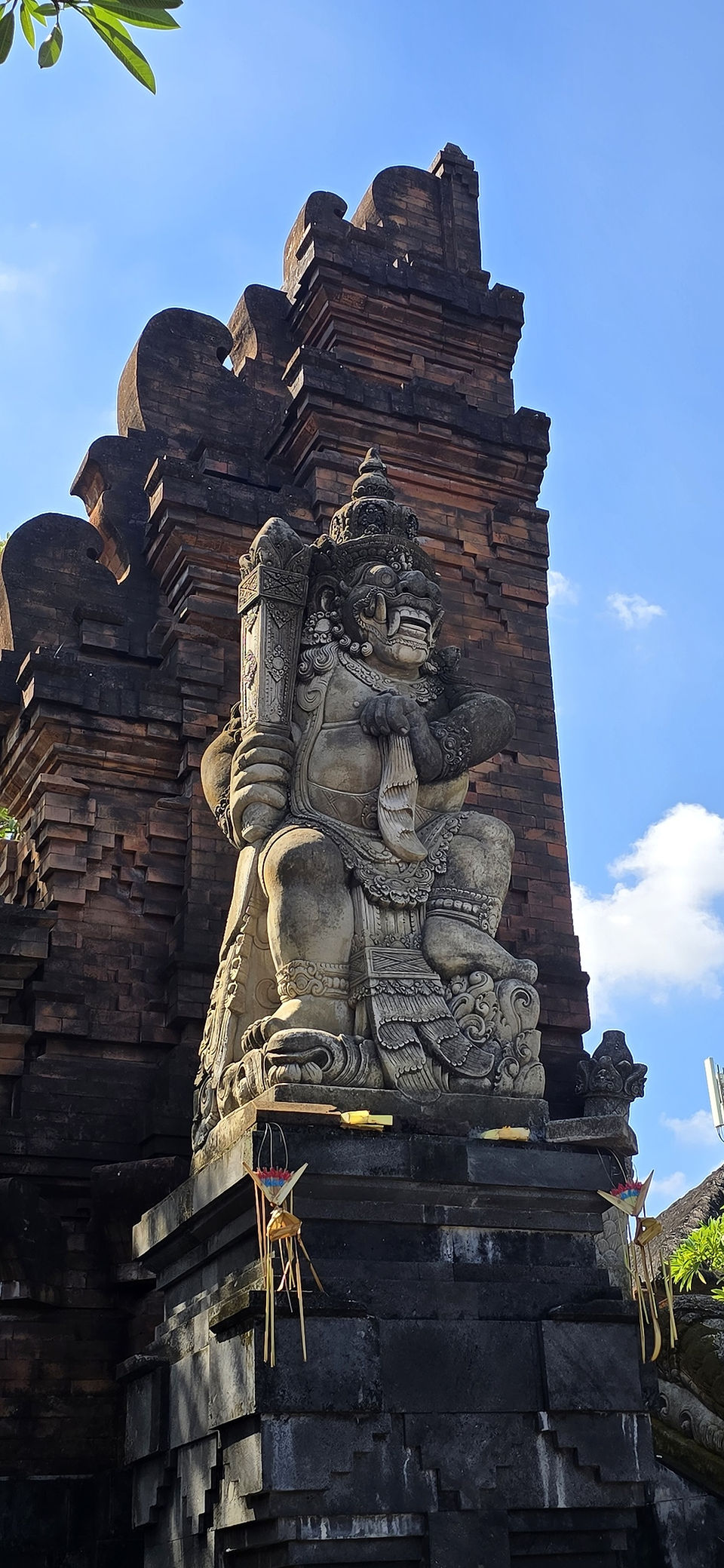 Stone guardian statue at a temple gate, holding a club, against a bright blue sky with offerings below.