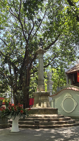 Tall stone pillar with a statue on top, set in a lush garden with a large tree. Nearby is a vase of red flowers and a building with a flag.