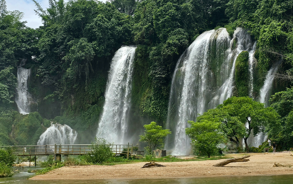 Lush green waterfall scene with multiple cascades. Footbridge and trees in foreground. Serene and natural atmosphere.