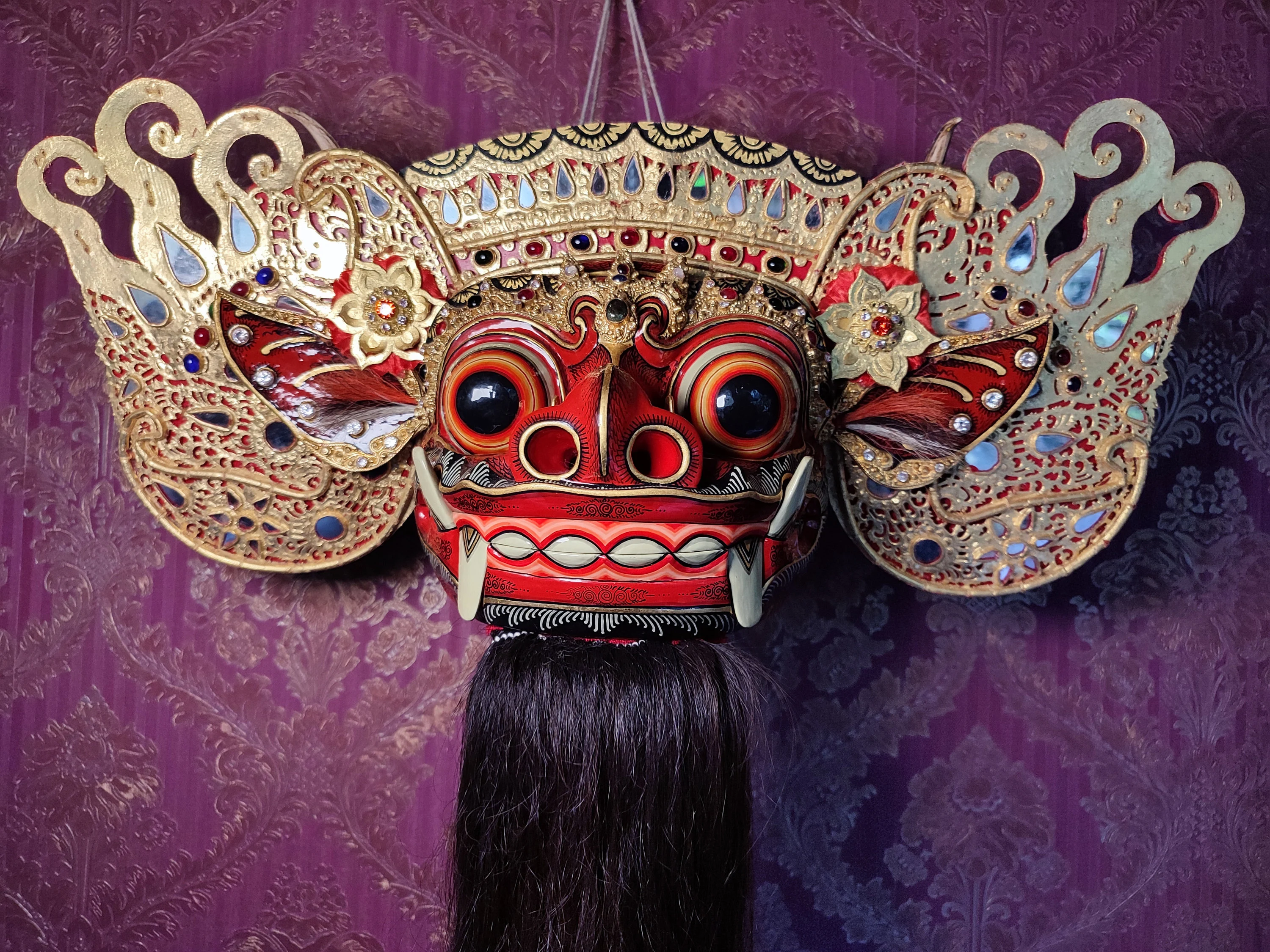 Intricate red and gold mask with large eyes, ornate patterns, and long black hair hangs against a purple, patterned background.