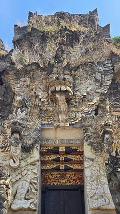 Ornate stone temple facade with giant carved face, red-gold door, overgrown plants on top, and clear blue sky overhead.