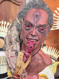 A detailed statue with a third eye and protruding tongue holds a decorated fan. The background features a pattern of white and brown hues.