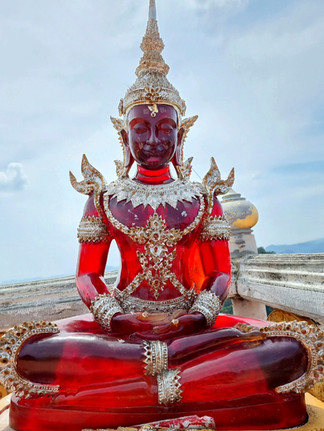 Red gemstone Buddha statue adorned with intricate silver patterns, seated in a meditative pose against a cloudy sky background.
