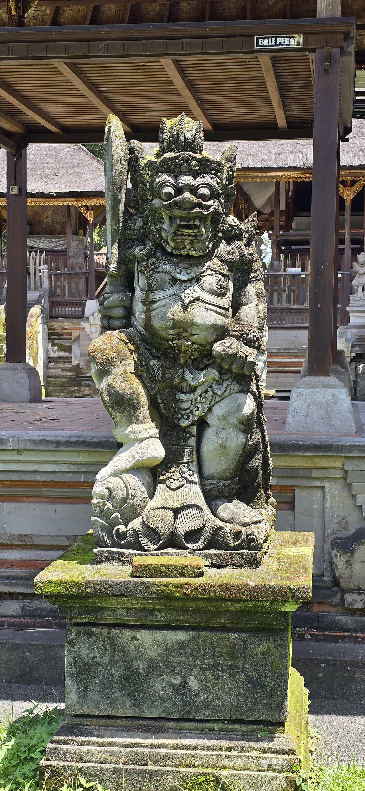 Stone Balinese guardian statue with moss, standing in front of a traditional wooden pavilion. Sign reads "BALE PEGAT."
