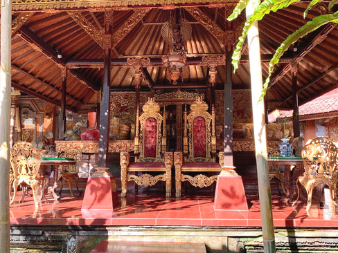 Ornate seating area with two red and gold thrones under a wooden pavilion. Intricate carvings and warm sunlight create a regal ambiance.