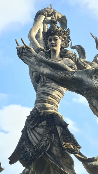 Ornate Balinese dedari statue with raised arms against a blue sky, featuring intricate carvings and flowing drapery.