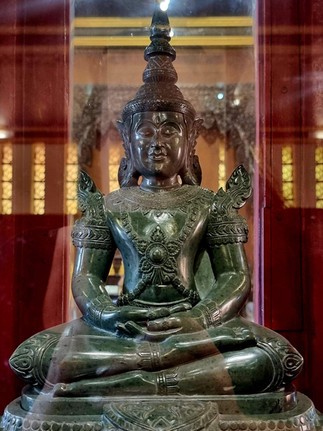 Intricately carved green statue of a seated figure in a serene pose inside a glass case, against a warm-toned, ornate background.