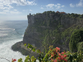 Cliffside temple with red rooftops overlooking a sparkling ocean. Blue sky, white clouds, and vibrant green foliage with pink flowers.