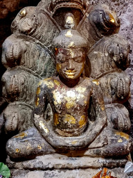 Ancient stone Buddha statue with gold leaf details, seated in meditation. Background features multi-headed serpent motifs. Earthy tones.