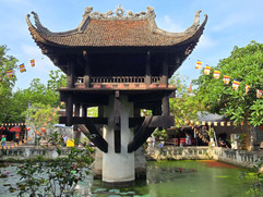 Pagoda on a pillar over a pond with lily pads, surrounded by trees and colourful flags. A few people in the background, vibrant setting.