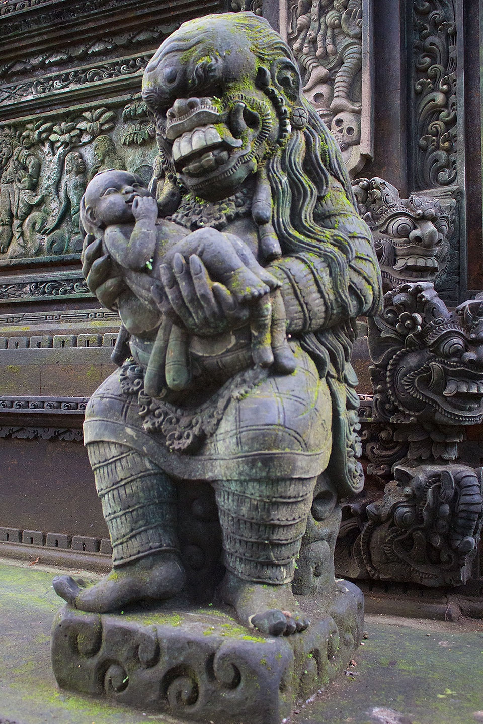 Detailed statue of Rangda holding a child, her face twisted in a fearsome grimace, moss enveloping the stone surface, standing sentinel at the cremation temple