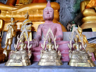 Multiple Buddha statues in various sizes and colors, with a large pink Buddha in the center. Gold hues dominate the serene setting.