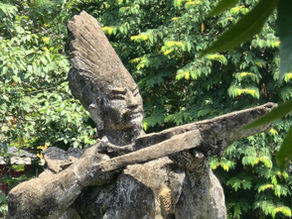 Stone statue of a figure playing a flute stands on a pedestal in a green pond, surrounded by lush trees and foliage in the background.