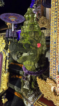 Ornate moss-covered stone statue with a pink flower in a temple, surrounded by colourful gold and purple decorations at night.