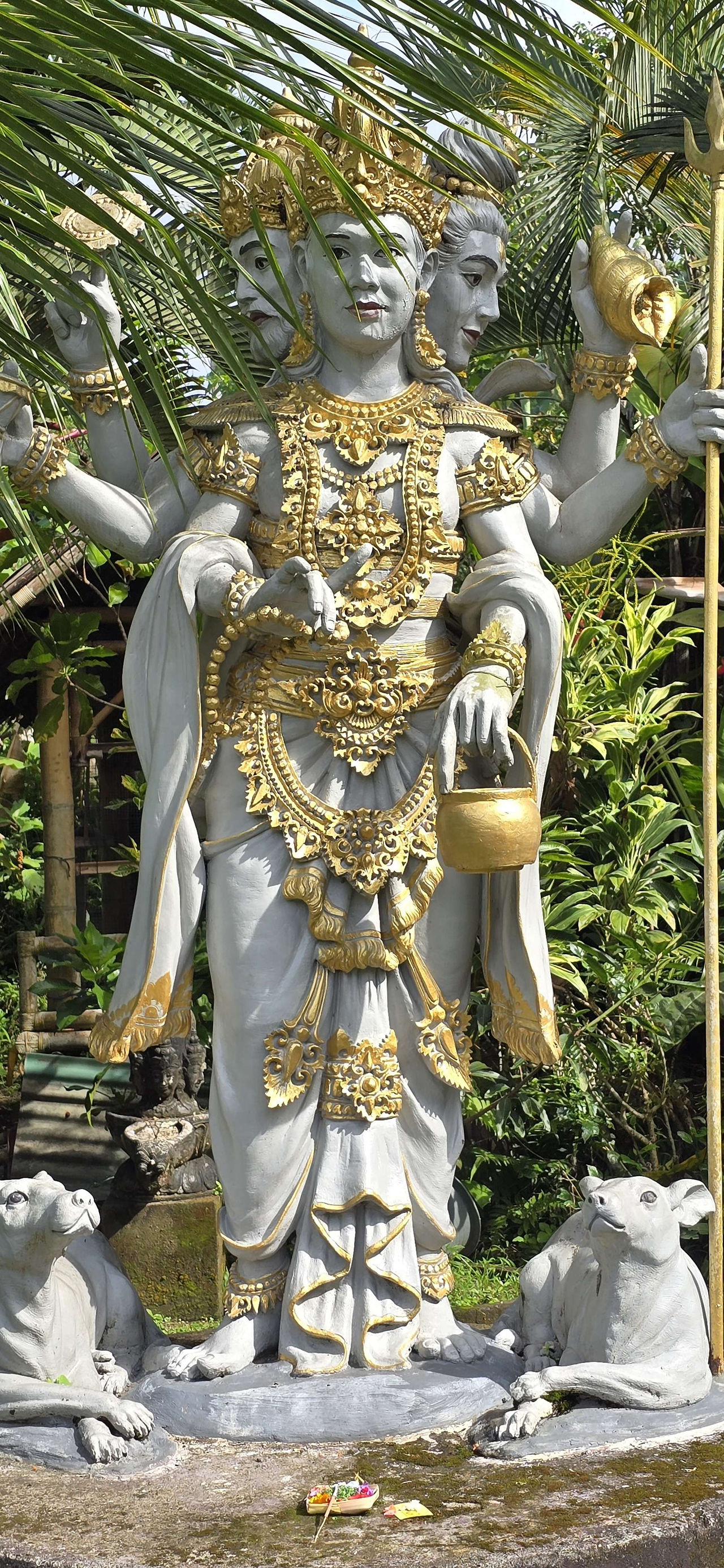Statue of a multi-armed deity adorned in gold, surrounded by greenery, with two stone dogs at the base. Sunlit, serene ambiance.