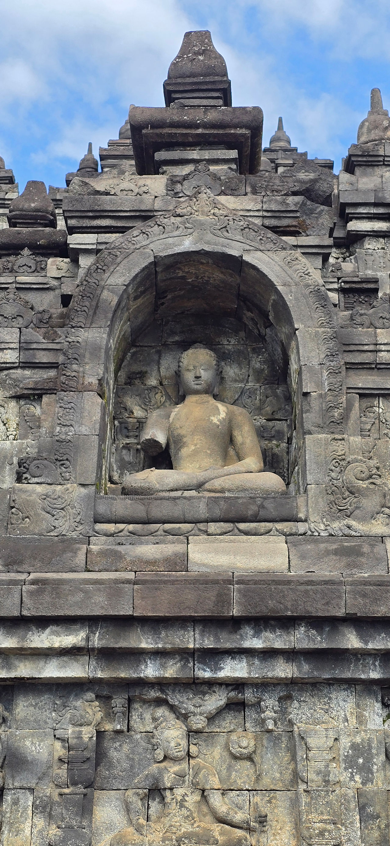 Stone Buddha statue in a niche on a temple wall, with intricate carvings below. Weathered stone, cloudy blue sky background.