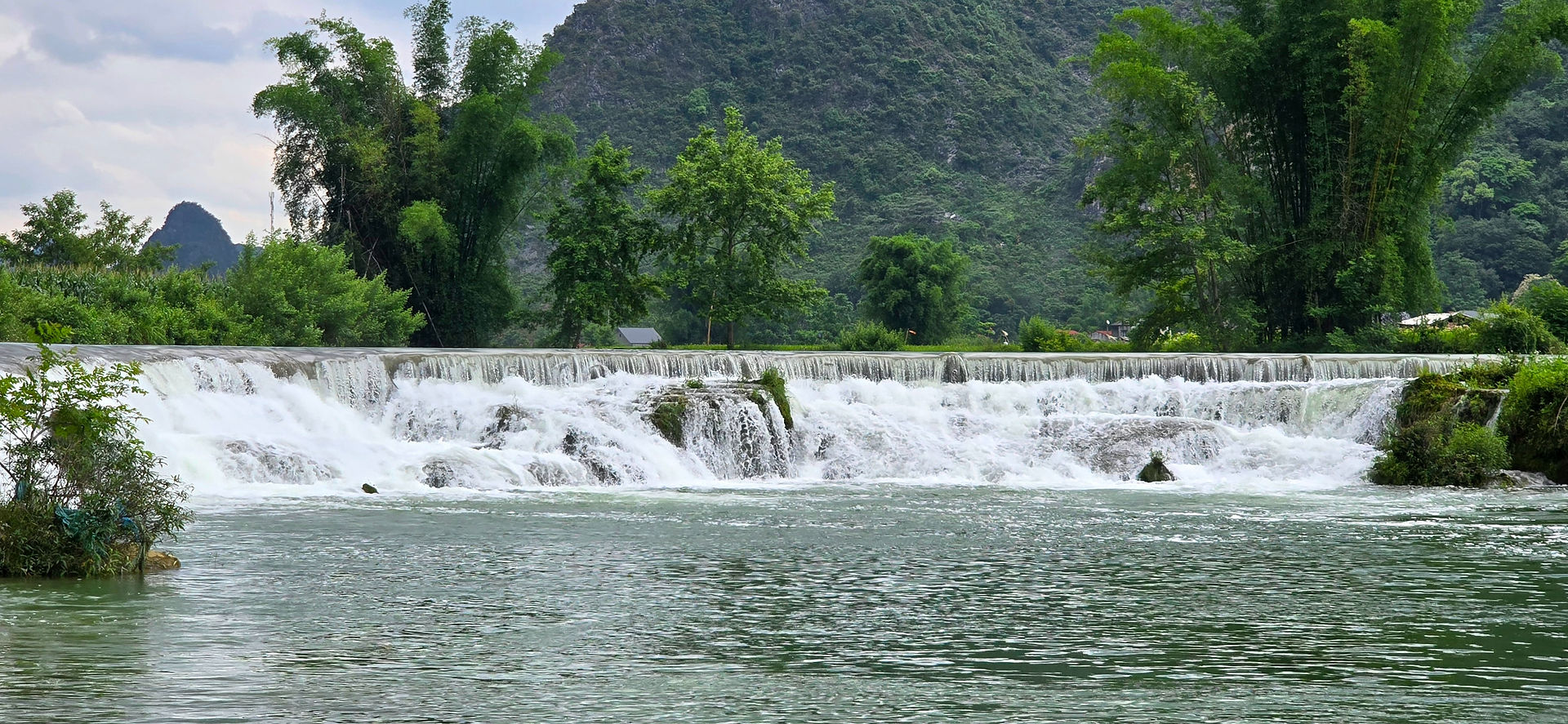 Peaceful landscape of flowing waterfall surrounded by lush greenery and distant mountains under a cloudy sky. Calm and serene mood.