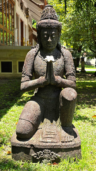 Stone statue of a kneeling figure with hands in prayer, holding a white flower. Surrounded by greenery, with a building in the background.