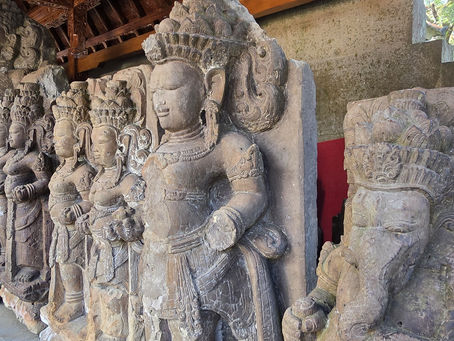 Row of weathered stone deity statues inside a temple pavilion, with carved crowns and serene faces under a wooden roof.