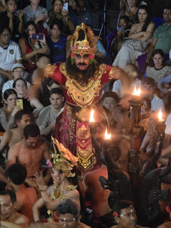 Performer in ornate costume dances with raised arms amid a seated crowd, lit by torches. Spectators watch, recording with phones.