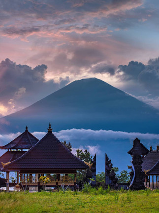 Sunrise over Mount Agung above Balinese temple roofs, pink clouds and misty blue hills.