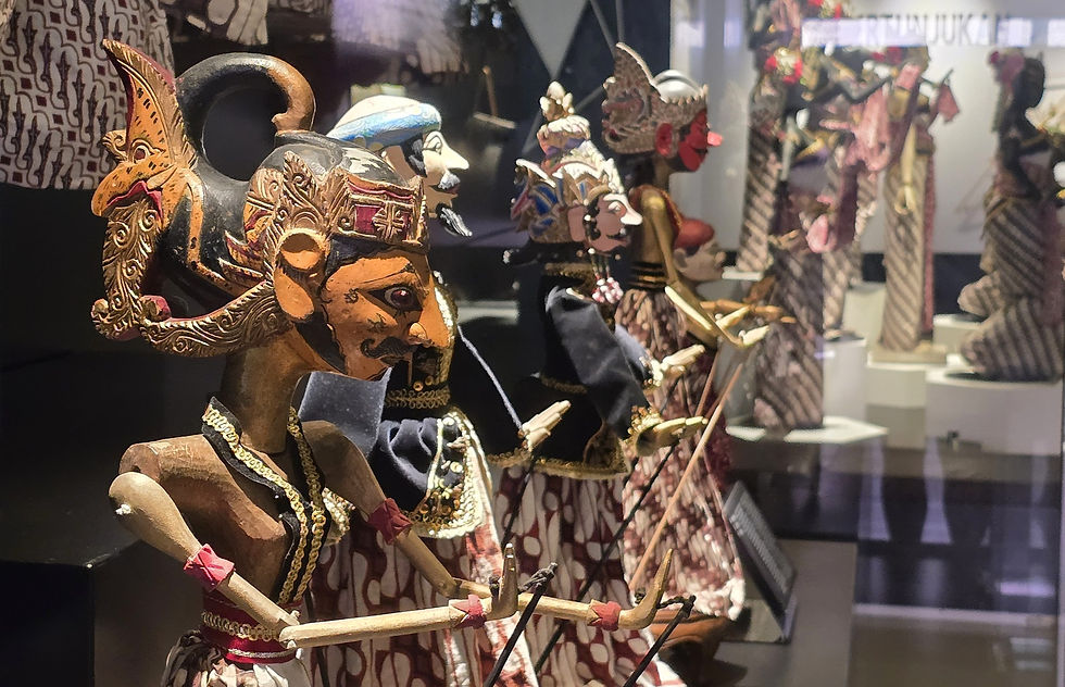 Colorful shadow puppets with intricate designs and patterns are displayed in a glass case. The background has a reflection of textiles.