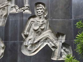 Stone relief of seated figure on textured wall, flanked by partial figures and green foliage. Gray tones dominate the scene.Historic stone gate surrounded by trees, colourful flags, and red flowers. Green park setting, creating a peaceful atmosphere.