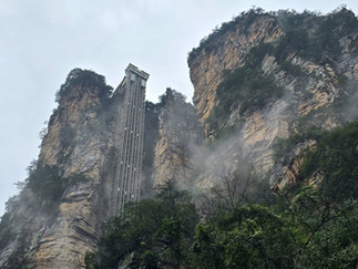 Tall glass elevator ascends between steep, misty cliffs with lush greenery, under a cloudy sky, creating a dramatic, ethereal scene.