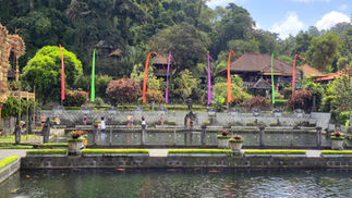 People relax near a stone pool, surrounded by lush greenery and brightly coloured flags in front of rustic houses under a blue sky.