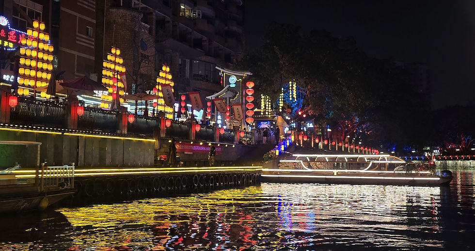 Night river scene with illuminated boats, colorful lanterns, and vibrant reflections on water. Bright neon signs and trees in the background.