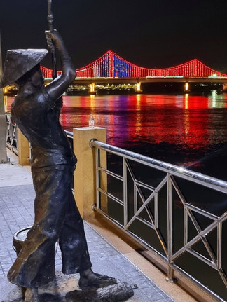 cast iron statue of a fisherman mid cast, illuminated red and blue bridge in the distance, reflecting widely in the river
