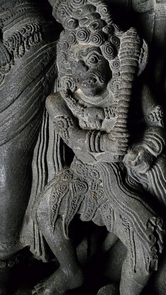 Intricate stone carving of a mythical figure holding a club. Detailed curls in hair and ornate clothing. Dark, textured background.