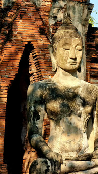 Ancient stone Buddha statue with serene expression in meditation pose, set against a backdrop of rustic red brick arches.
