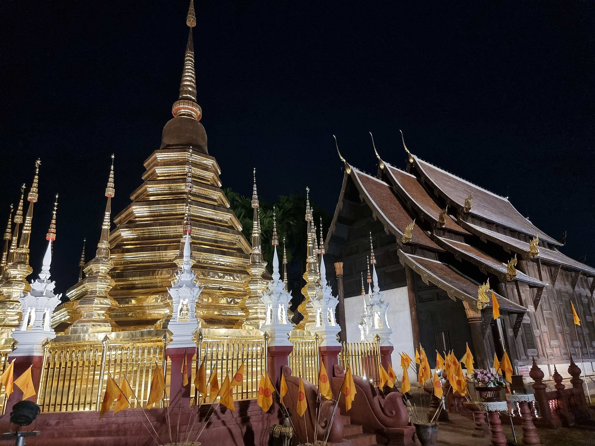 Golden spire adorns ornate temple at night, surrounded by orange flags and intricate details, set against a dark sky. Peaceful ambiance.