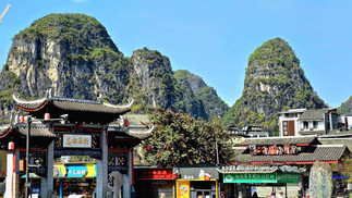 Traditional Chinese buildings with ornate roofs in front of lush green karst mountains under a clear blue sky. Signs in Chinese are visible.
