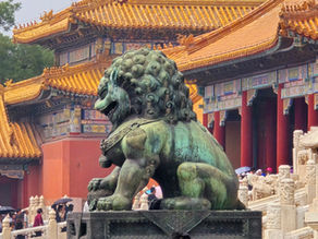 Bronze lion statue in front of ornate, golden-roofed building with detailed patterns. Tourists with umbrellas in the foreground.