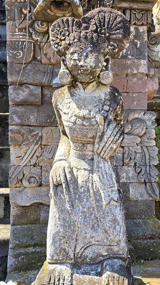 Weathered stone statue of a seated woman with ornate hair and jewelry, in front of carved temple stonework, sunlit.