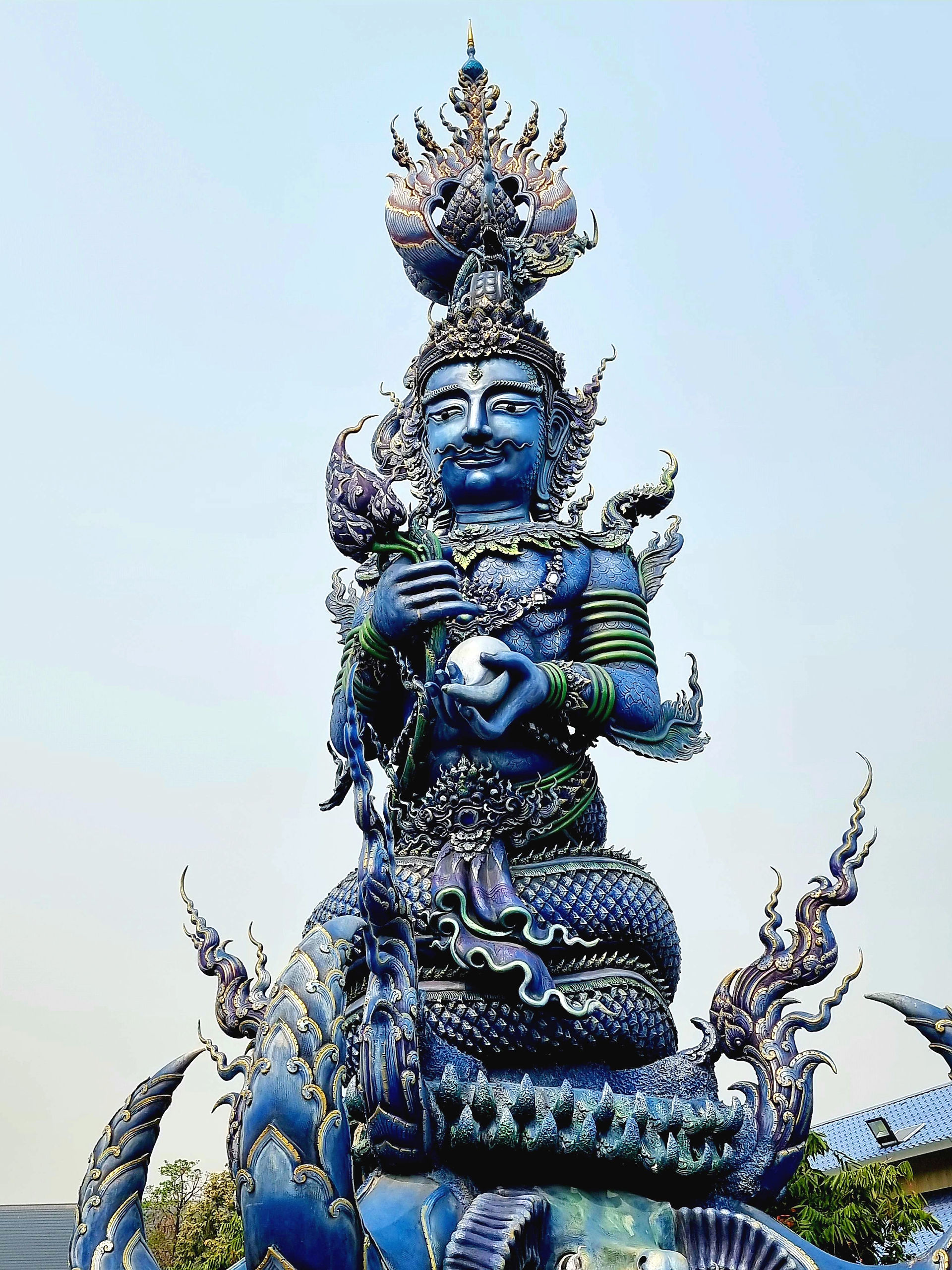 A towering blue deity-like statue with a serpentine lower body, holding a glowing pearl and a lotus staff. The figure’s head is encircled by ornate flames and crowned with a lotus finial, standing on a coiled naga base