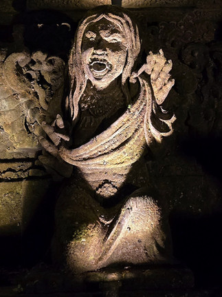 Stone sculpture of a woman with flowing hair, expressive face, and raised arms, illuminated dramatically against a dark, detailed background.