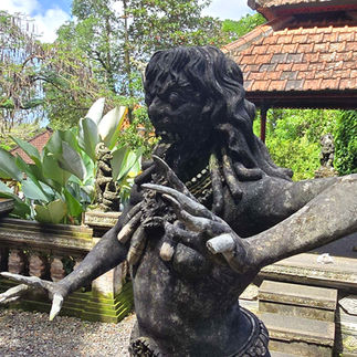 Stone statue of a demonic mythical figure with outstretched arms in a lush garden setting. Background includes tropical plants and a tiled pavilion.