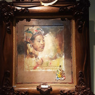 Ornate wooden frame with painted portrait of a woman, colourful headscarf, script below. Golden decorative text at the top. Warm tones.