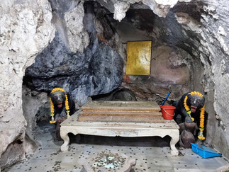Two stone panthers with garlands guard a cave altar. Coins scatter the floor; a red bucket and blue dustpan are beside the altar.