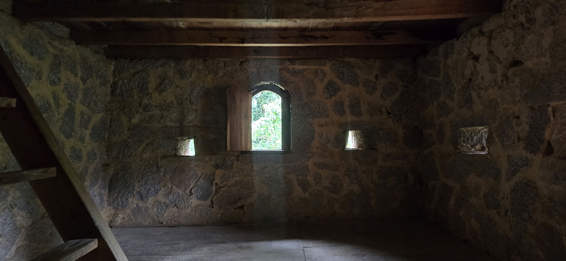 Stone-walled room with a wooden ceiling, a beam of light through an arched window. Stairs on the left; serene mood, greenery outside.